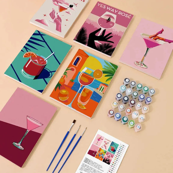 Cocktail-themed paint-by-numbers set, featuring six ready-made 20x15 cm paintings, paint pots, three brushes, and instructions arranged on the table.