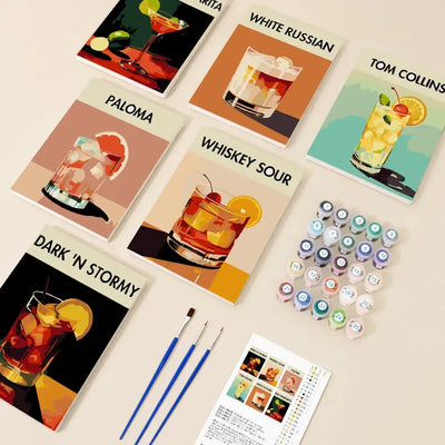 Classic Cocktails paint by numbers set, featuring six 20x15 cm drink-themed paintings with names, paint pots, brushes, and instructions arranged on the table.