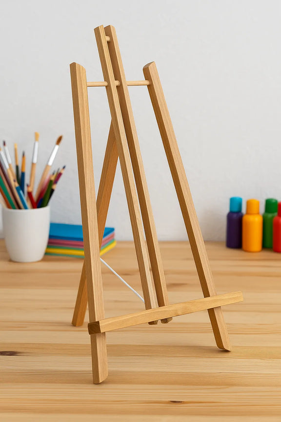 A lightweight wooden easel on the craft table. The easel is about 50 cm high and 27 cm wide, perfect for table use in paintings and paint-by-numbers. In the background, brushes and colorful painting supplies can be seen.
