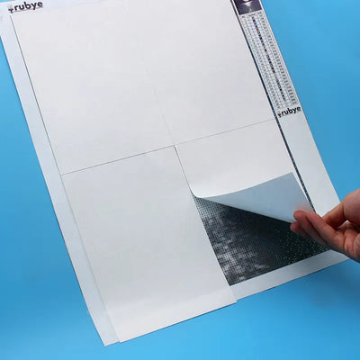 In the image, a hand lifts the corner of the protective sheet from the diamond painting.